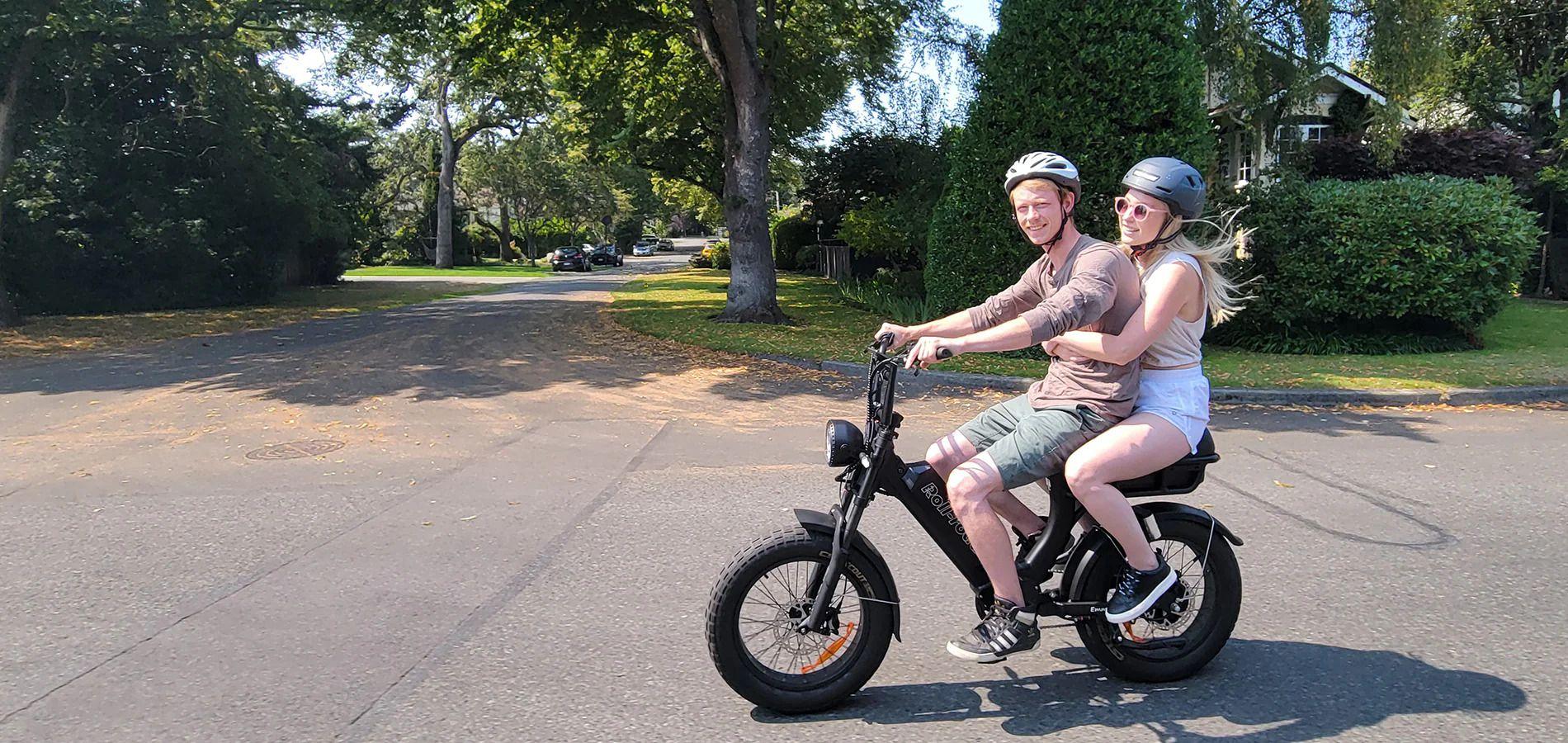 Two people riding an electric bike on a sunny day in a residential neighborhood with trees and greenery.