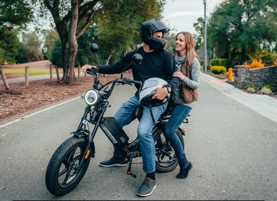 Two people sitting on a motorcycle parked on a paved path surrounded by trees and greenery.